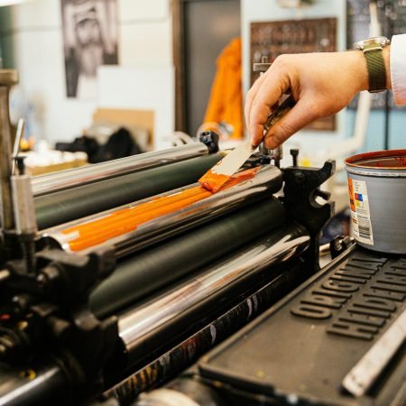 A detailed view of a hand applying orange ink to a vintage printing press roller indoors.